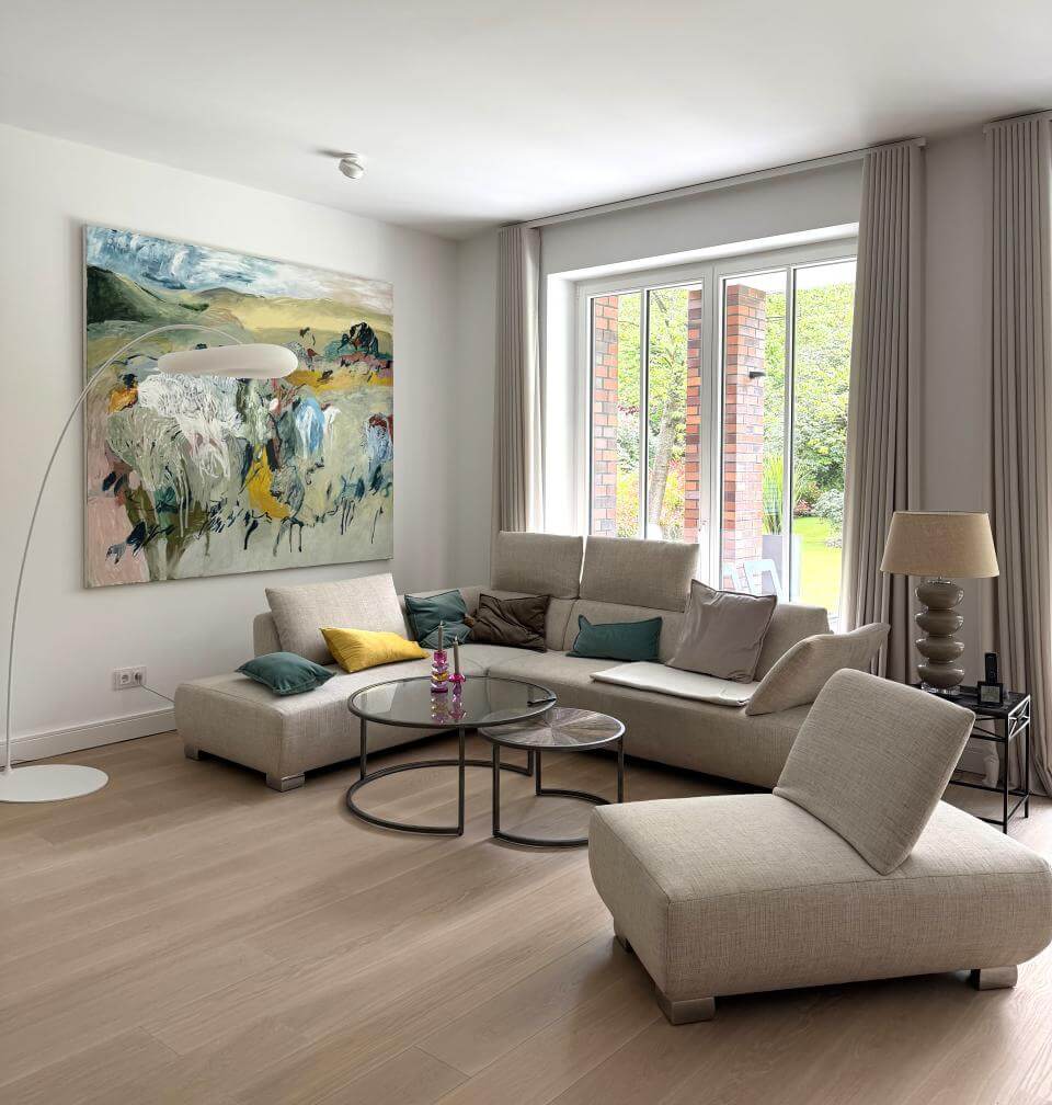 Modern eingerichtetes Wohnzimmer mit hellem Ecksofa, Glastisch und großem abstrakten Gemälde von Hans Sieverding, viel Tageslicht durch bodentiefe Fenster und Blick in den Garten