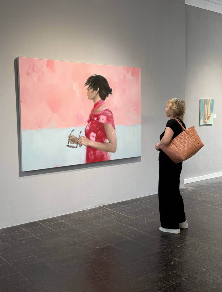 Ausstellungsszene in der Galerie RIECK: Besuchende betrachten ein großformatiges, zeitgenössisches Porträtgemälde in Rosa‑ und Pastelltönen im modernen Ausstellungsraum.