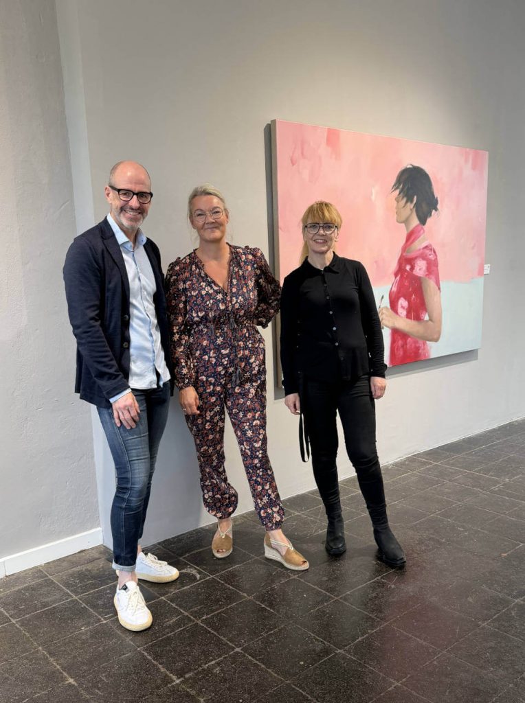 Ausstellungsszene in einer modernen Galerie: die Galerieinhaber der Galerie RIECK und die Künstlerin Siri Gindesgaard stehen vor einem großformatigen, zeitgenössischen Porträtgemälde in Rosa‑ und Pastelltönen und erleben Kunst im gemeinsamen Dialog.
