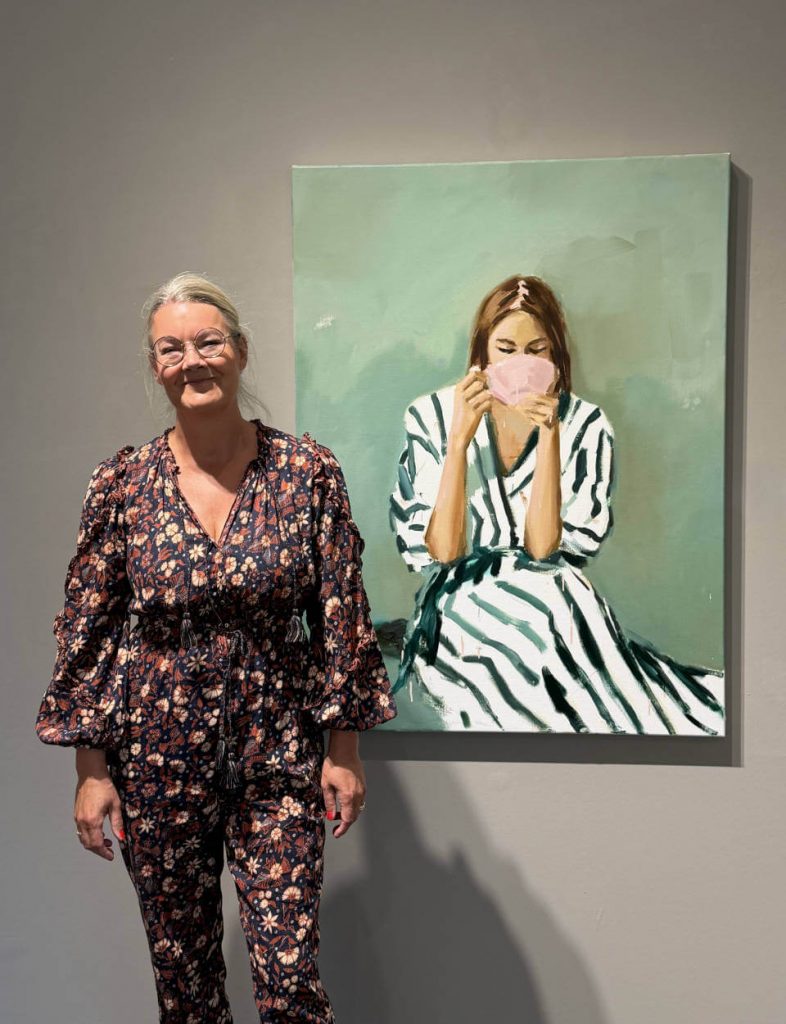 Galerieszene mit der Künstlerin Siri Gindesgaard vor einem zeitgenössischen Porträtgemälde in Grün‑ und Weißtönen – moderner Ausstellungsraum, Kunst im direkten Dialog zwischen Werk und Publikum.