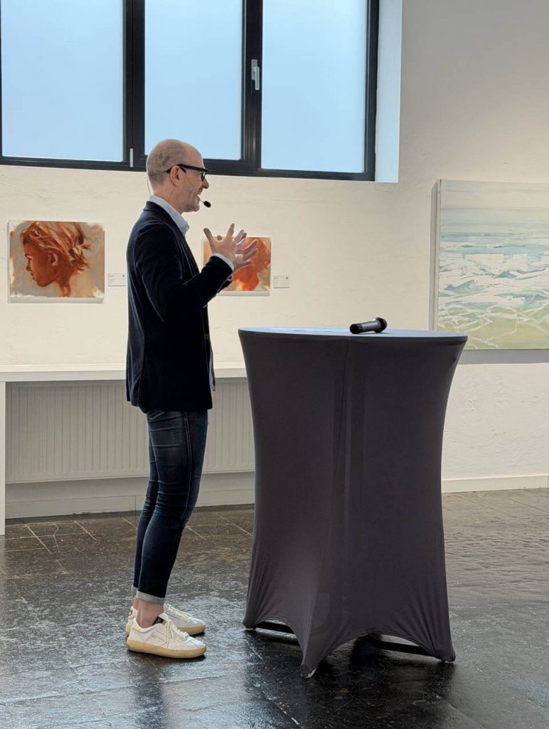 Galerieeröffnung mit Vortragssituation: Galerieinhaber der Galerie RIECK spricht am Stehtisch vor zeitgenössischen Gemälden in hellem Ausstellungsraum – lebendige Kunst‑ und Ausstellungsszene.