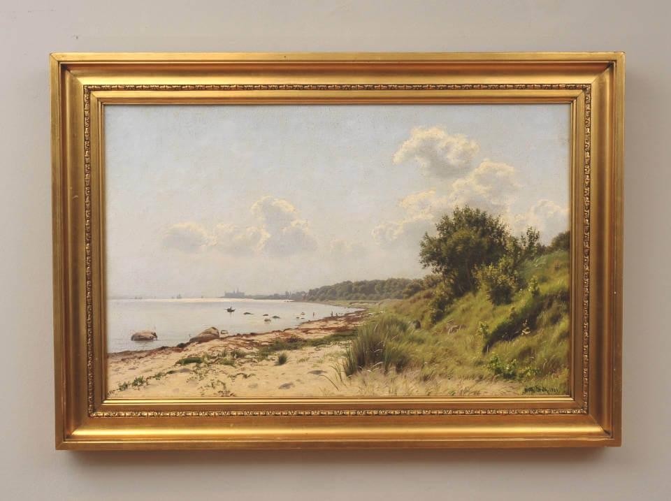 Landschaftsgemälde einer ruhigen Küste mit Sandstrand, vereinzelten Steinen und flachem Wasser, gesäumt von grünen Uferpflanzen und Bäumen unter hellem, leicht bewölktem Himmel. Gerahmt in einem historischen Goldrahmen. Von Vilhelm Groth bei Galerie RIECK.
